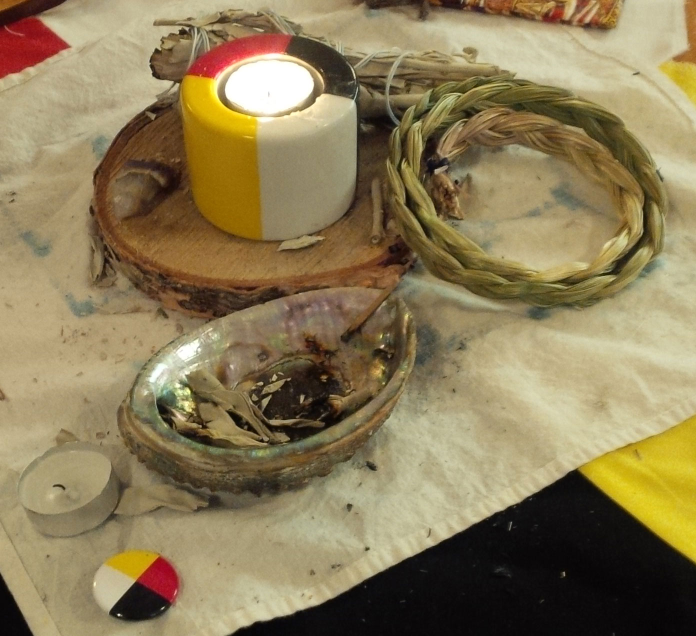 four directions candle lit with sweet grass strands laying around it on a cloth on top of a four directions flag